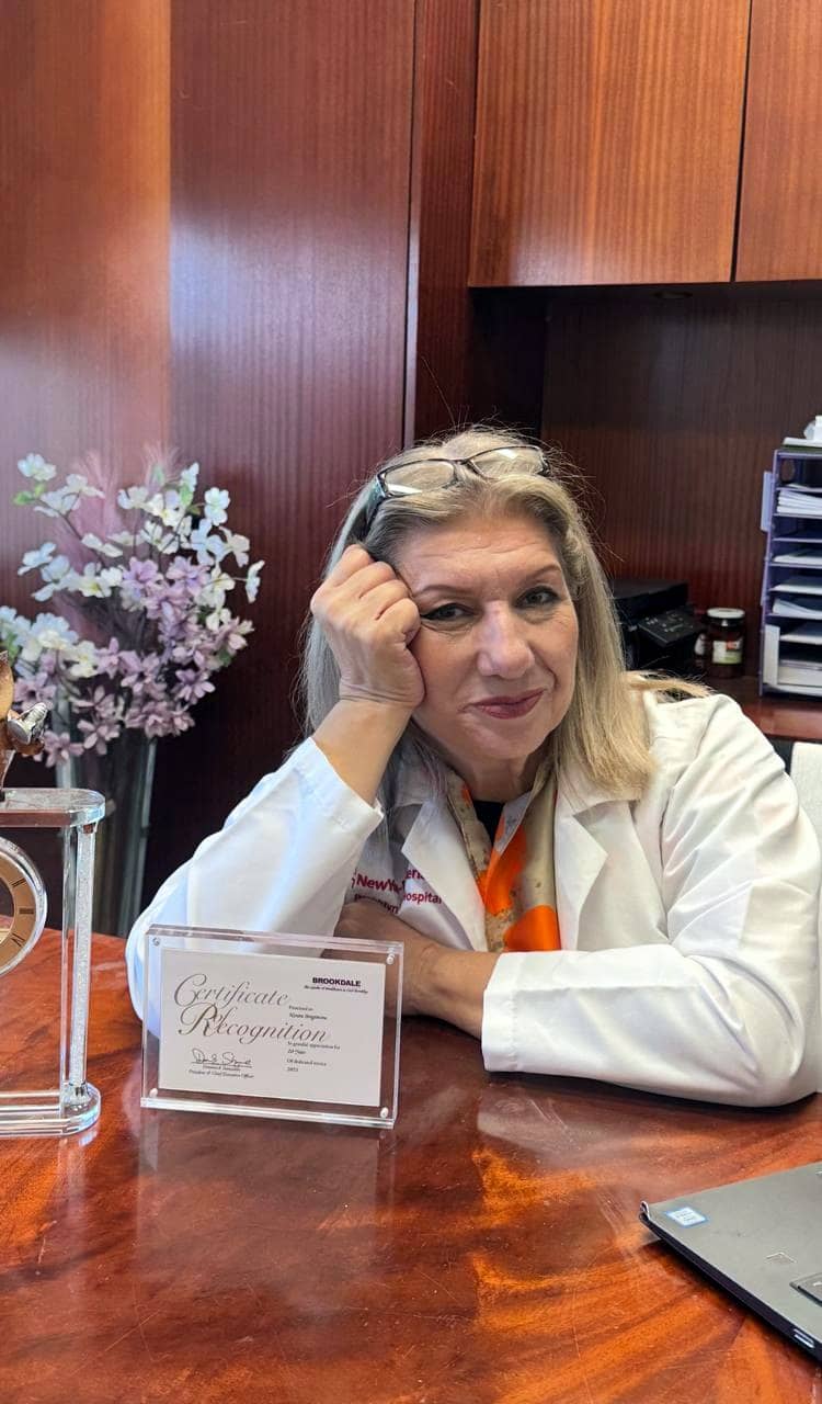 Ninara Ibragimova in a white coat at a desk, a certificate of recognition and a clock in front of her