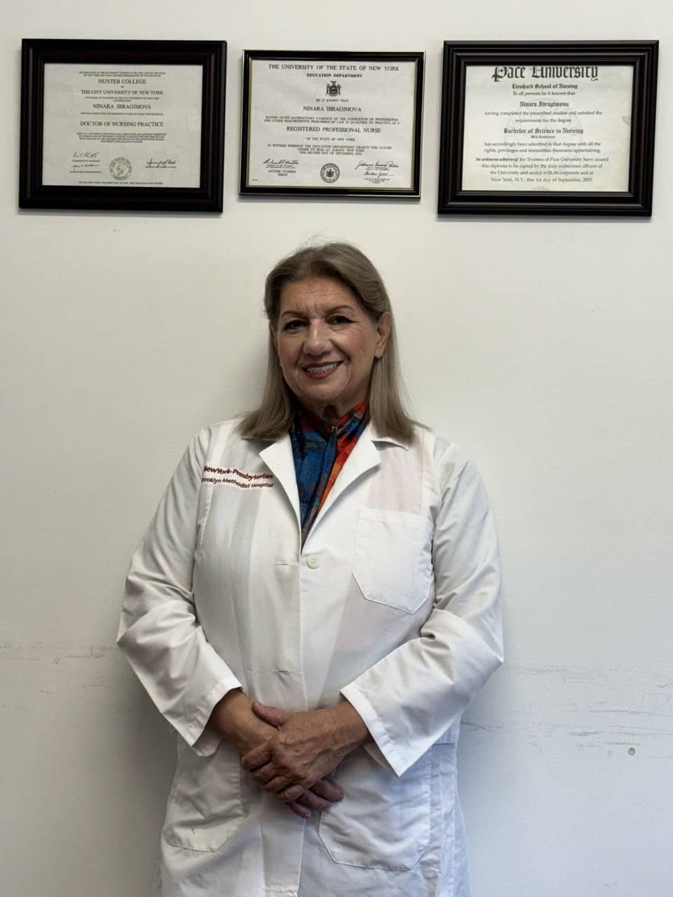 Нинара Ибрагимова в белом халате улыбается на фоне дипломов Hunter College, штата Нью-Йорк и Pace University
